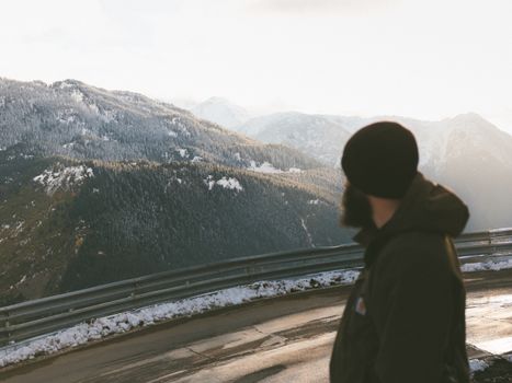 Side view unrecognizable bearded man in warm clothes standing on serpentine road and admiring rough forested hills covered with snow