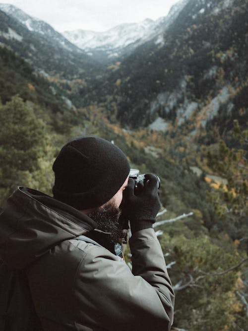 Side view anonymous bearded male hiker in outerwear taking photos on professional photo camera of rough forested mountains covered with snow in daylight