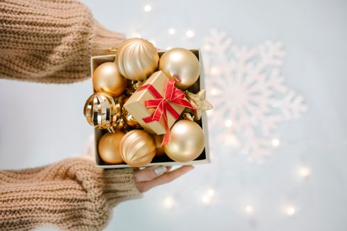 Free From above of crop anonymous female in warm sweater demonstrating box with Christmas baubles against white background with glowing garland Stock Photo