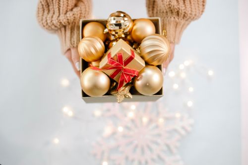 Free From above of crop anonymous woman in sweater showing box with golden Christmas baubles against light background with glowing garland Stock Photo