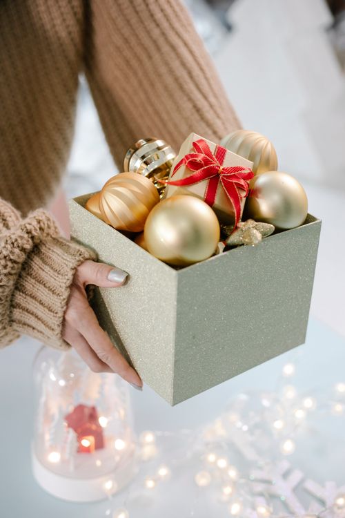 Free Crop unrecognizable female in warm sweater showing box with stylish golden Christmas baubles in light room Stock Photo