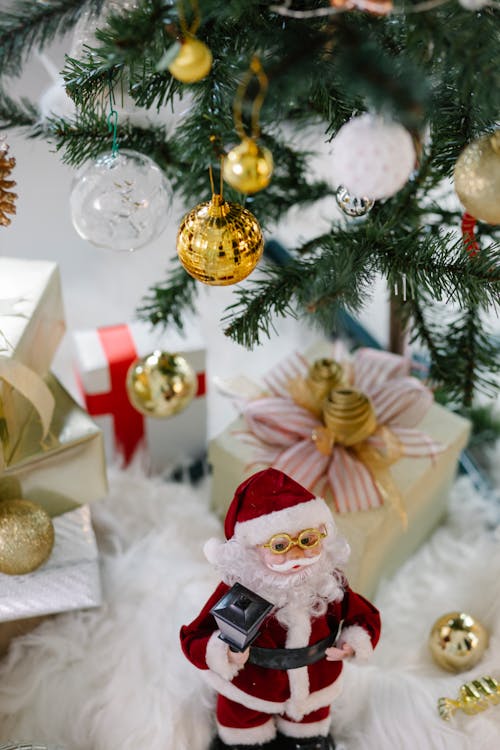 Free From above of small toy Santa Claus placed among boxes of presents under Christmas tree with colorful baubles in light apartment Stock Photo