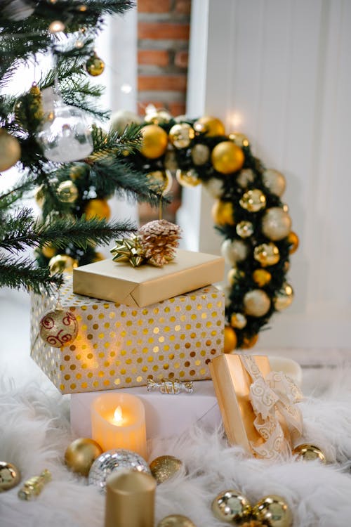 Present boxes wrapped in colorful decorative paper placed near wreath and baubles with candle under coniferous Christmas tree