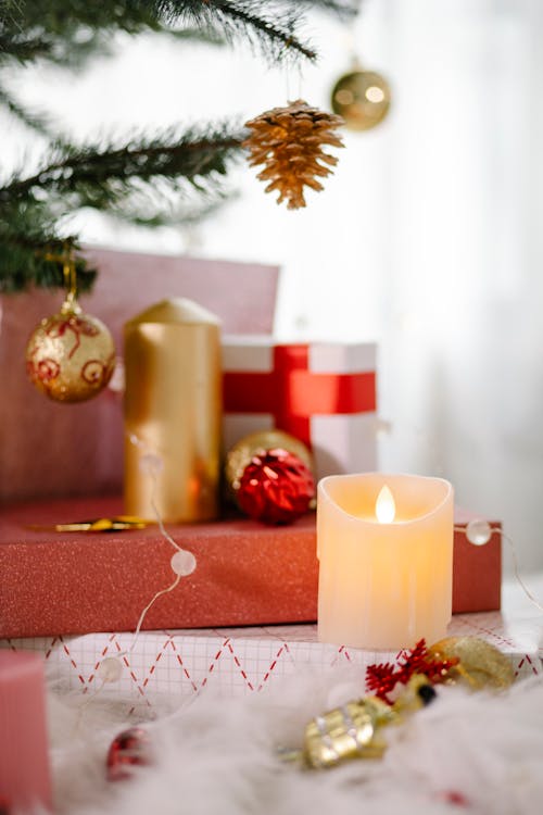 Burning candle and pile of present boxes wrapped in decorative paper placed under Christmas tree decorated with cones and baubles