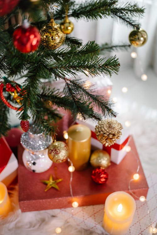 From above of bright red present box with candles and fairy lights placed under Christmas tree decorated with baubles and cones