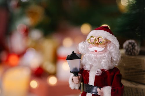 Small toy of Santa Claus in glasses with lantern near gift boxes on blurred background