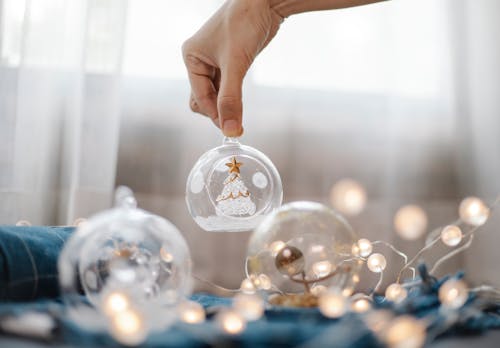 Crop anonymous person showing glass ball with decorative tree figure under shining garland on plaid