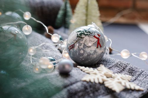 Free Christmas decorative snowflake near ball painted with bear in hat and scarf near garland on baggy textile near small fake fir placed on dark surface in bright place Stock Photo