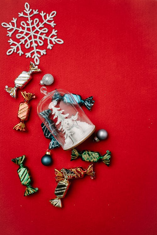 Top view of candies in colorful foils and small baubles scattered on red table with decorative snowflake and Christmas toy