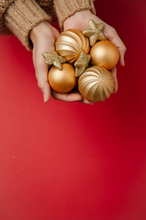 Free From above of crop unrecognizable female in warm sweater demonstrating pile of golden baubles and star shaped toys during Christmas preparation against red background Stock Photo