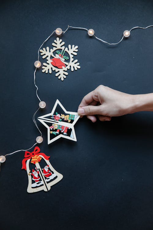 From above of crop unrecognizable lady showing creative wooden Christmas tree toys in shapes of star bell and snowflake against black background with glowing garland