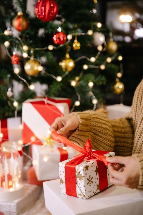 Free Crop anonymous female in warm sweater unpacking gift box decorated with red ribbon bow while sitting near Christmas tree during holidays Stock Photo