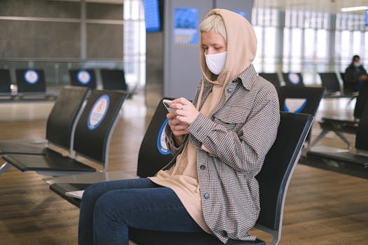 A woman wearing a face mask and a hood sits in an airport waiting area, using her smartphone.