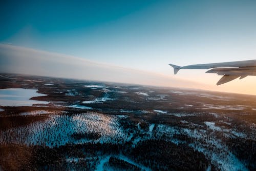 Through window of wing of modern aircraft flying over snowy terrain with lush trees and frozen lake at sunrise