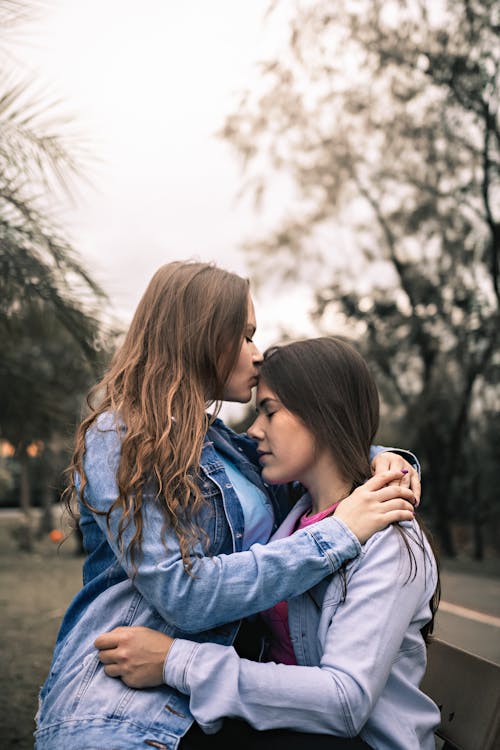 Free Side view of young homosexual woman in casual clothes sitting on knees of girlfriend and kissing forehead while resting together in park on autumn day Stock Photo