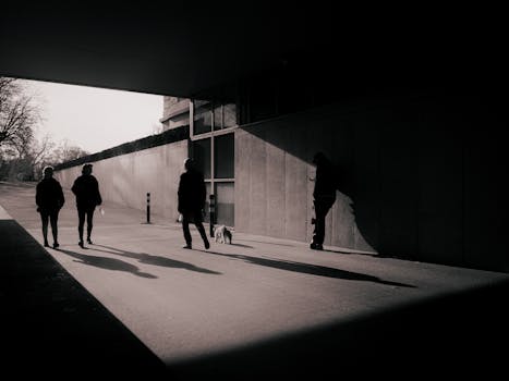 Monochrome scene with silhouetted figures walking under a bridge, casting long shadows.