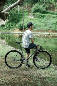 Side view full body of male bicyclist in cap sitting on bicycle on grassy coast near calm river in nature