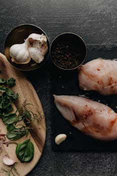 Dark moody photo of raw chicken breasts with garlic, spices, and fresh herbs on a slate surface.
