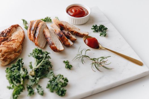 Sliced grilled chicken with greens and ketchup on a marble board, perfect for a healthy meal.
