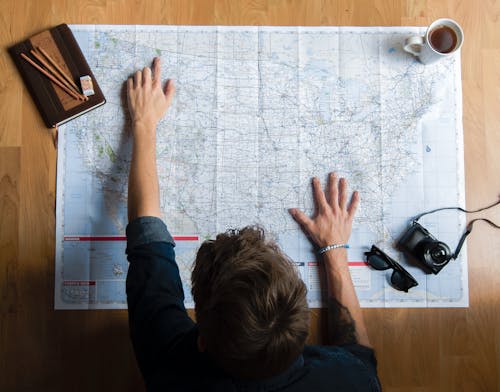 Free Top view of anonymous young male in casual clothes sitting on wooden floor with photo camera and coffee cup and pointing on paper map while planning trip route Stock Photo