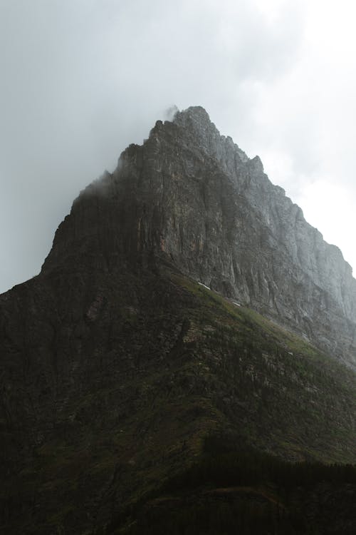 From below of huge rocky mountain with grassy slope and sharp peak hidden under foggy sky