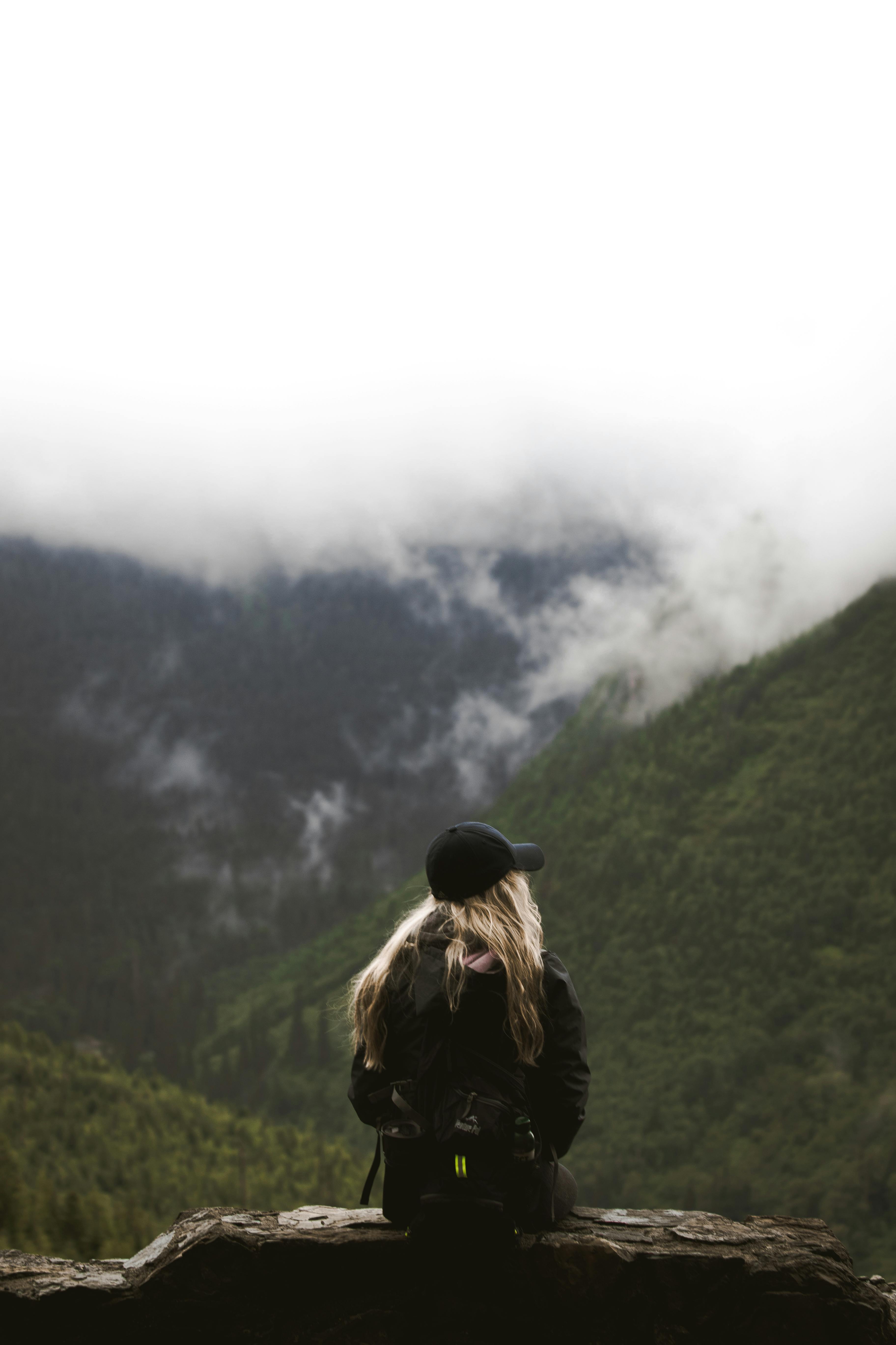 Back view of anonymous female explorer with long blond hair in casual outfit and cap sitting on edge of viewpoint and observing spectacular landscape of green mountains against cloudy sky