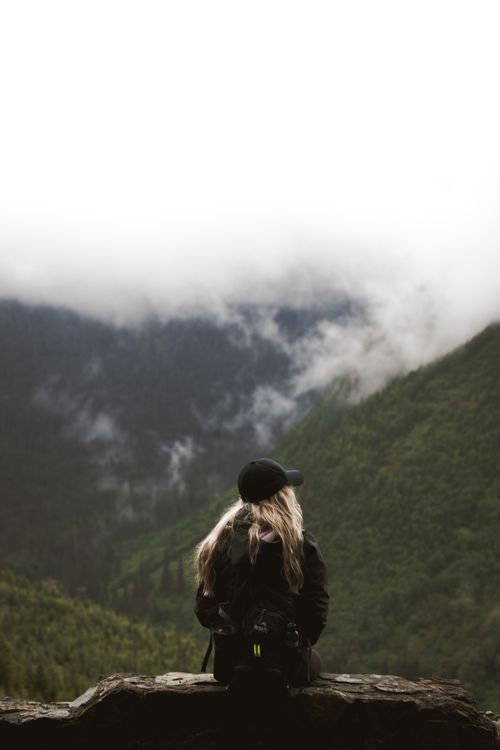 Free Back view of anonymous female explorer with long blond hair in casual outfit and cap sitting on edge of viewpoint and observing spectacular landscape of green mountains against cloudy sky Stock Photo