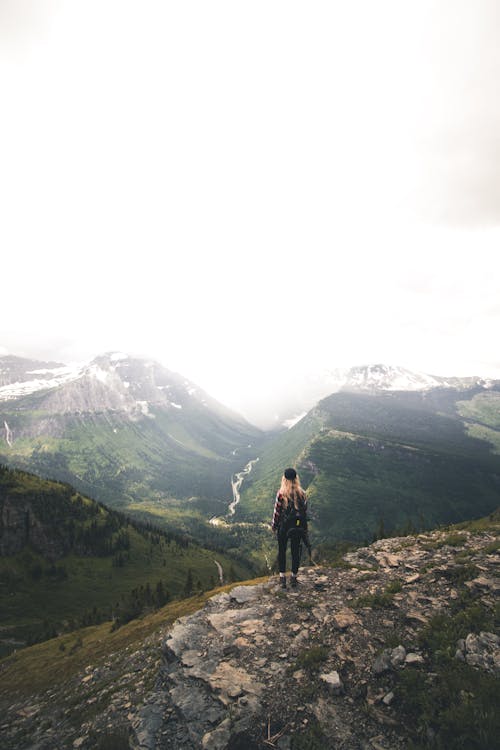 Back view of unrecognizable young female hiker with long blond hair in casual clothes and backpack recreating on rocky hill slope and admiring spectacular mountain view on cloudy day