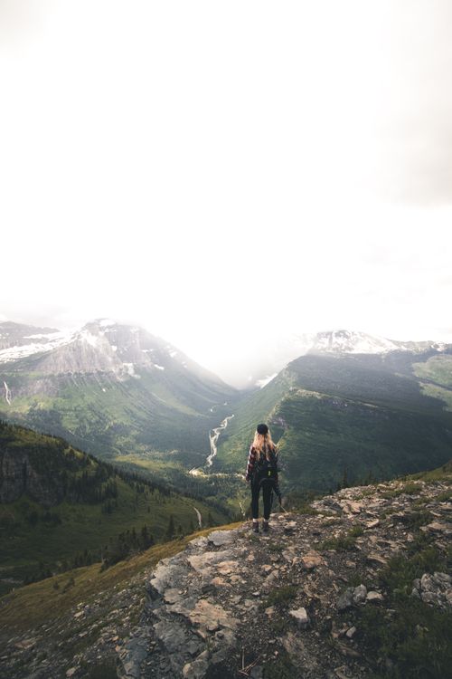 Free Back view of unrecognizable young female hiker with long blond hair in casual clothes and backpack recreating on rocky hill slope and admiring spectacular mountain view on cloudy day Stock Photo