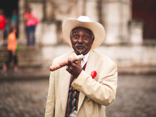 Free Senior surprised ethnic male entrepreneur in suit and hat showing cigar while looking at camera in town Stock Photo