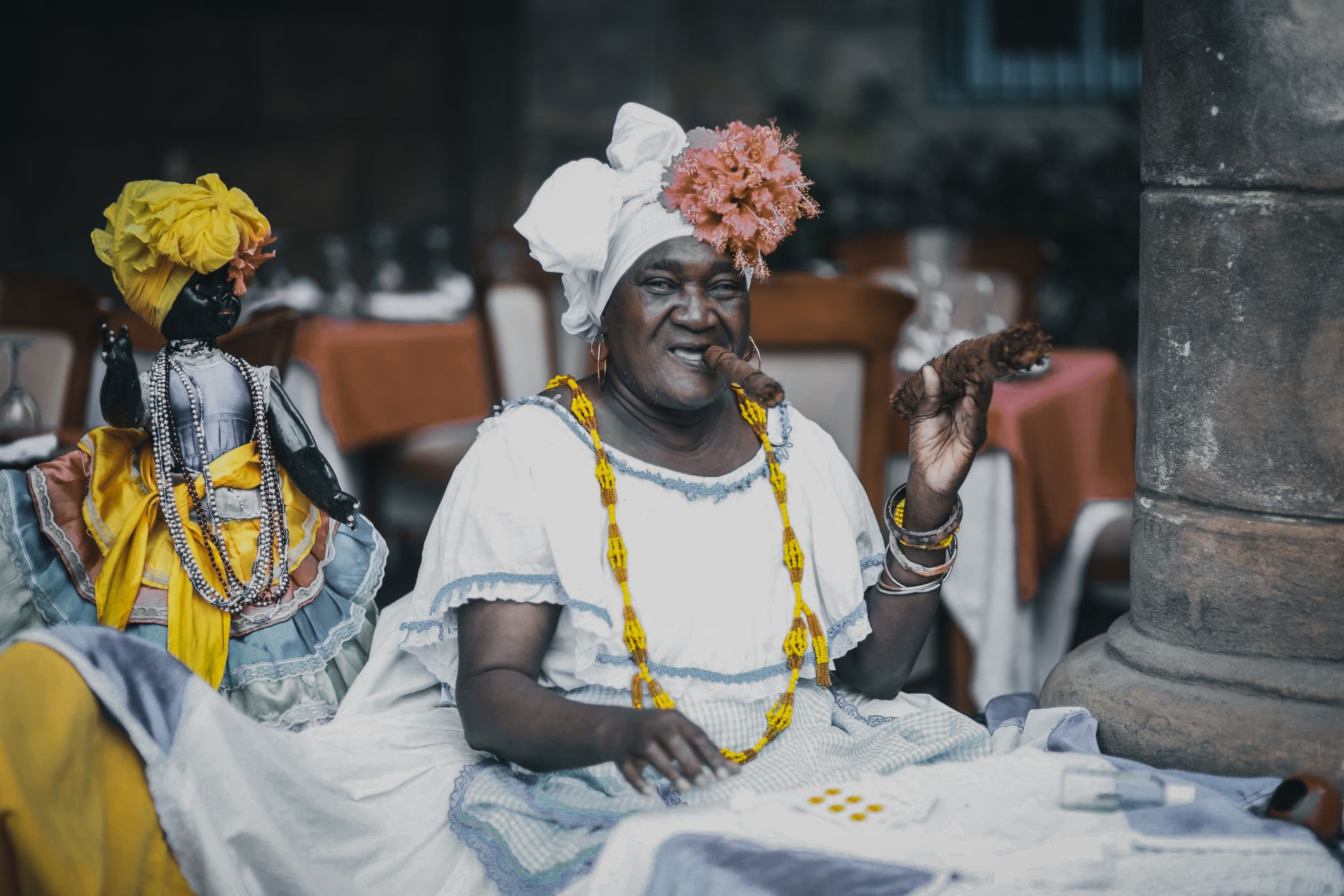 Content senior ethnic female in traditional apparel with cigar looking at camera near black doll with beads and headscarf