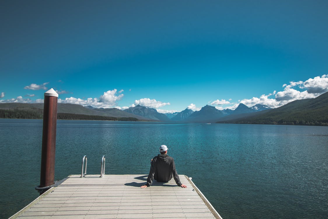 Back view of unrecognizable young male tourist in casual clothes and cap relaxing on wooden pier and admiring picturesque view of lake surrounded by rocky mountains against bright blue sky