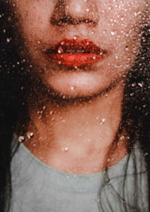 Crop unrecognizable young lady face view through glass with rain drops wearing red lipstick and casual outfit
