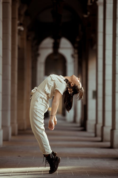 Free Side view of full body of female dancer wearing white overall standing in graceful posture among columns Stock Photo