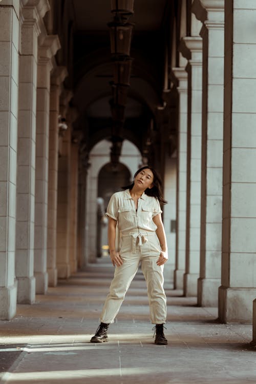 Full body of stylish provocative ethnic female in overall standing between aged building columns