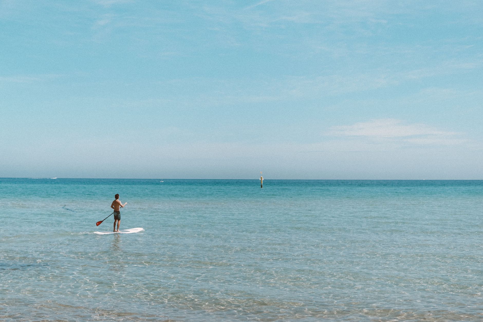 Un hombre solo haciendo paddle surf en un océano azul claro bajo un cielo soleado, exudando tranquilidad.