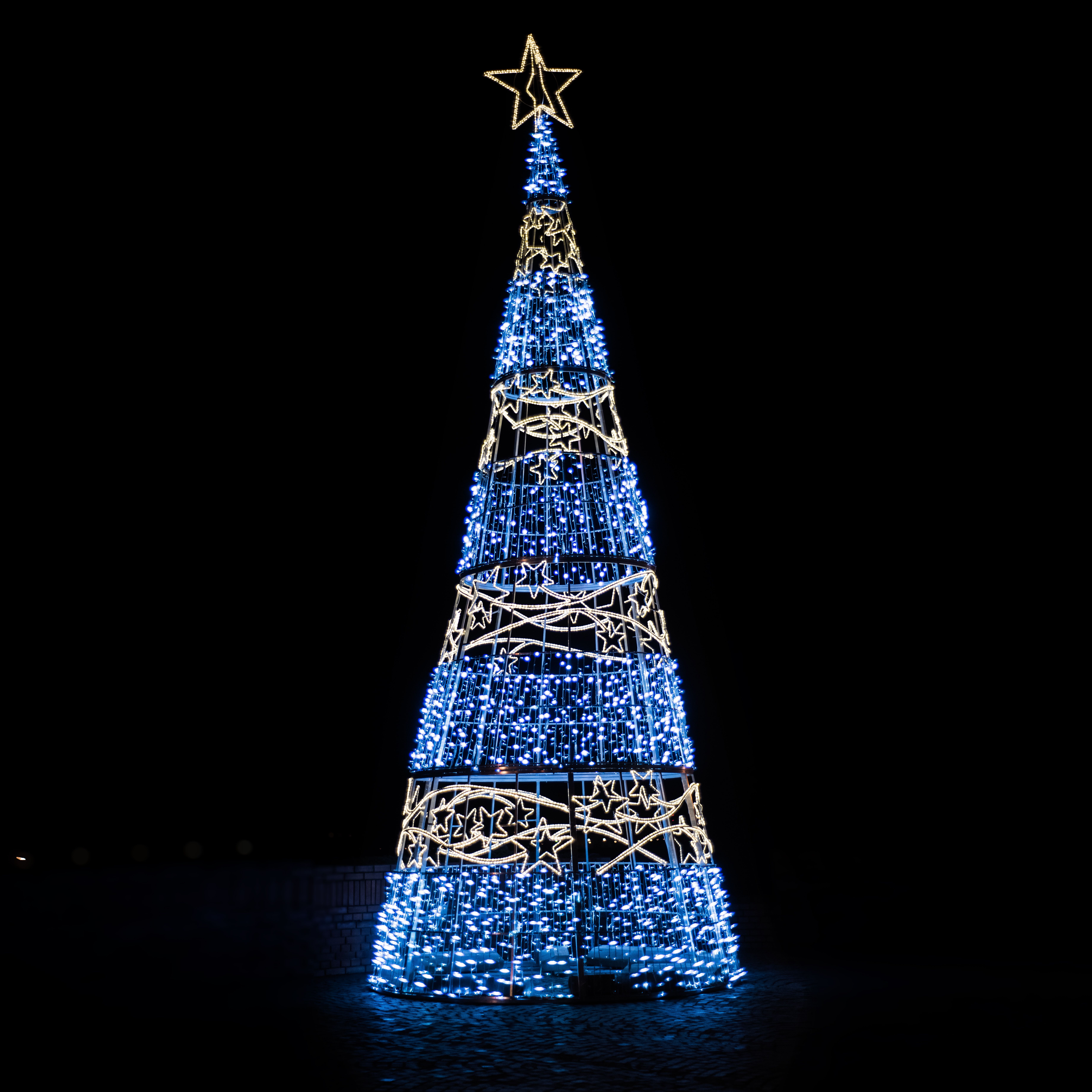 Illuminated Christmas tree with glowing golden and blue garlands topped with decorative star placed on black background during holiday celebration