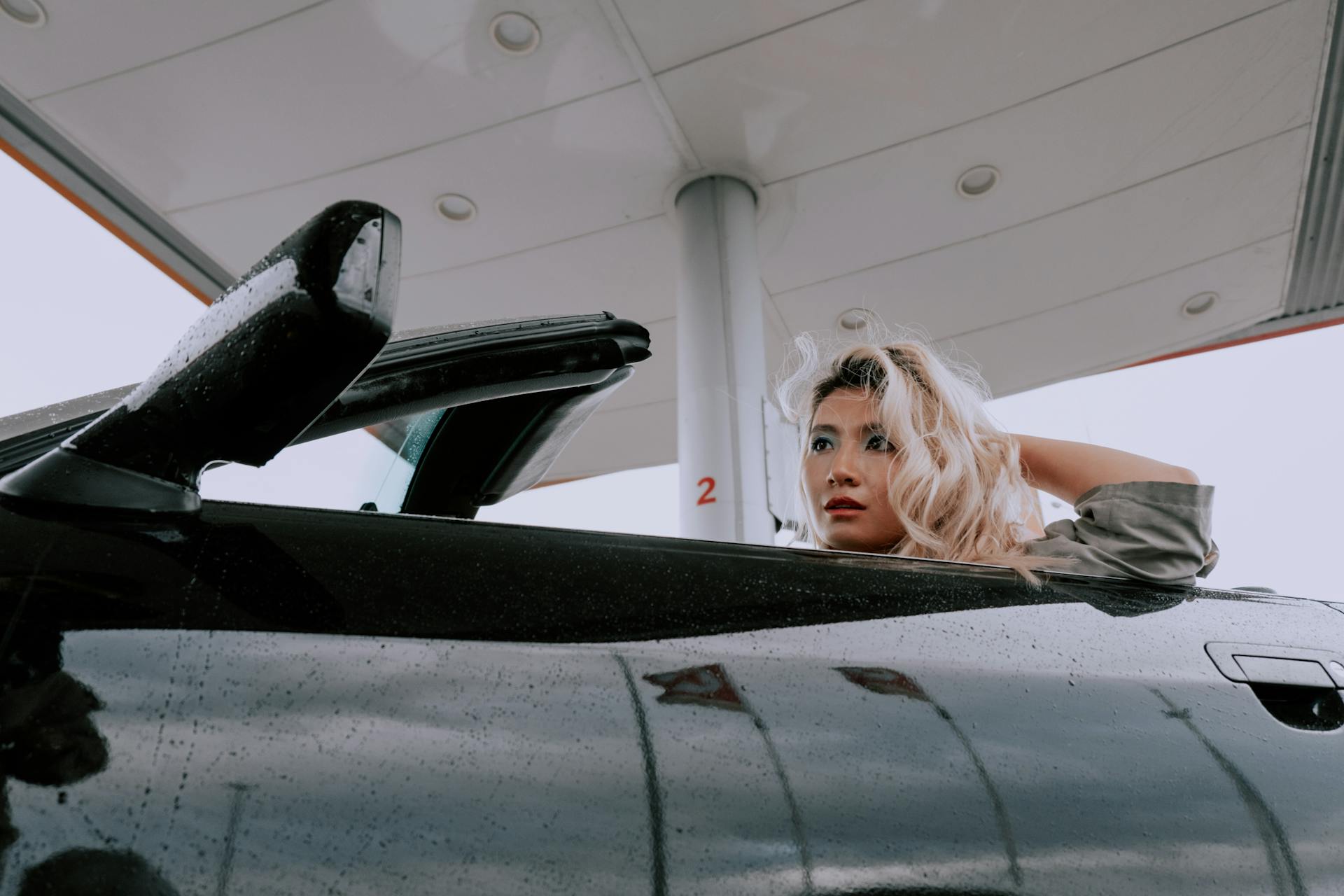 A fashionable woman sits in a convertible at a modern gas station, captured from a low angle.