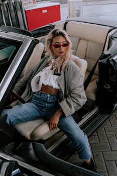 Fashionable woman in jeans and sunglasses sitting in convertible car outdoors.