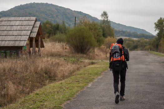 Back view full body of anonymous hiker with backpack strolling on asphalt roadway near shabby house inn suburb area during trekking
