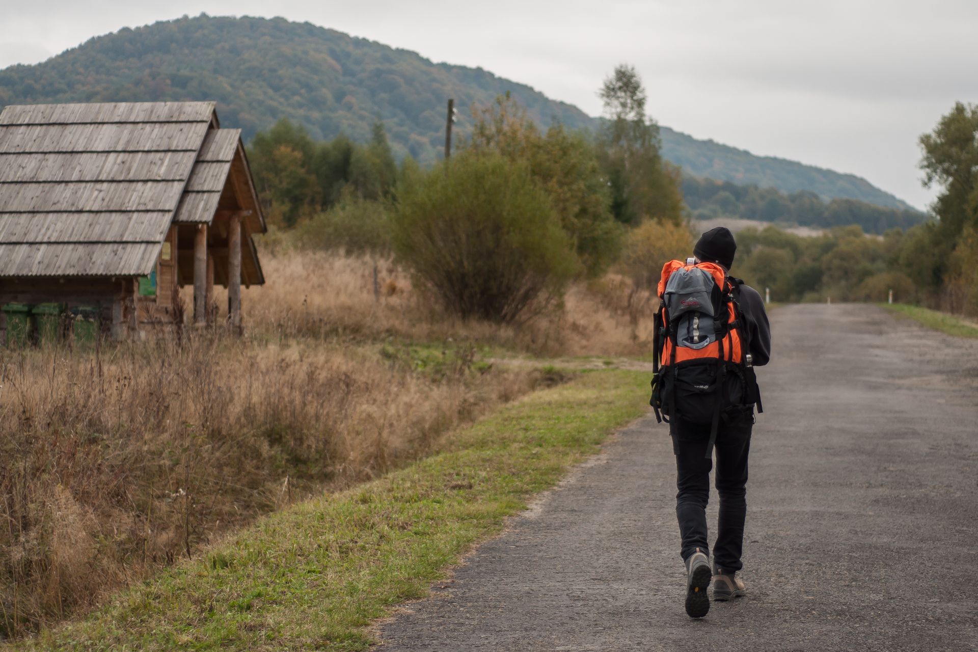 A lone backpacker walks on a rural path beside a wooden hut in Podkarpackie, Poland, surrounded by nature.