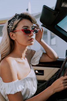 Stylish woman in sunglasses and headscarf driving convertible on a sunny day.