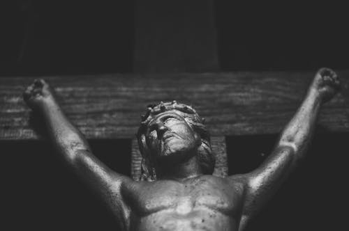 Black and white religious aged statue of Jesus Christ crucified on shabby wooden cross located in church against black wall