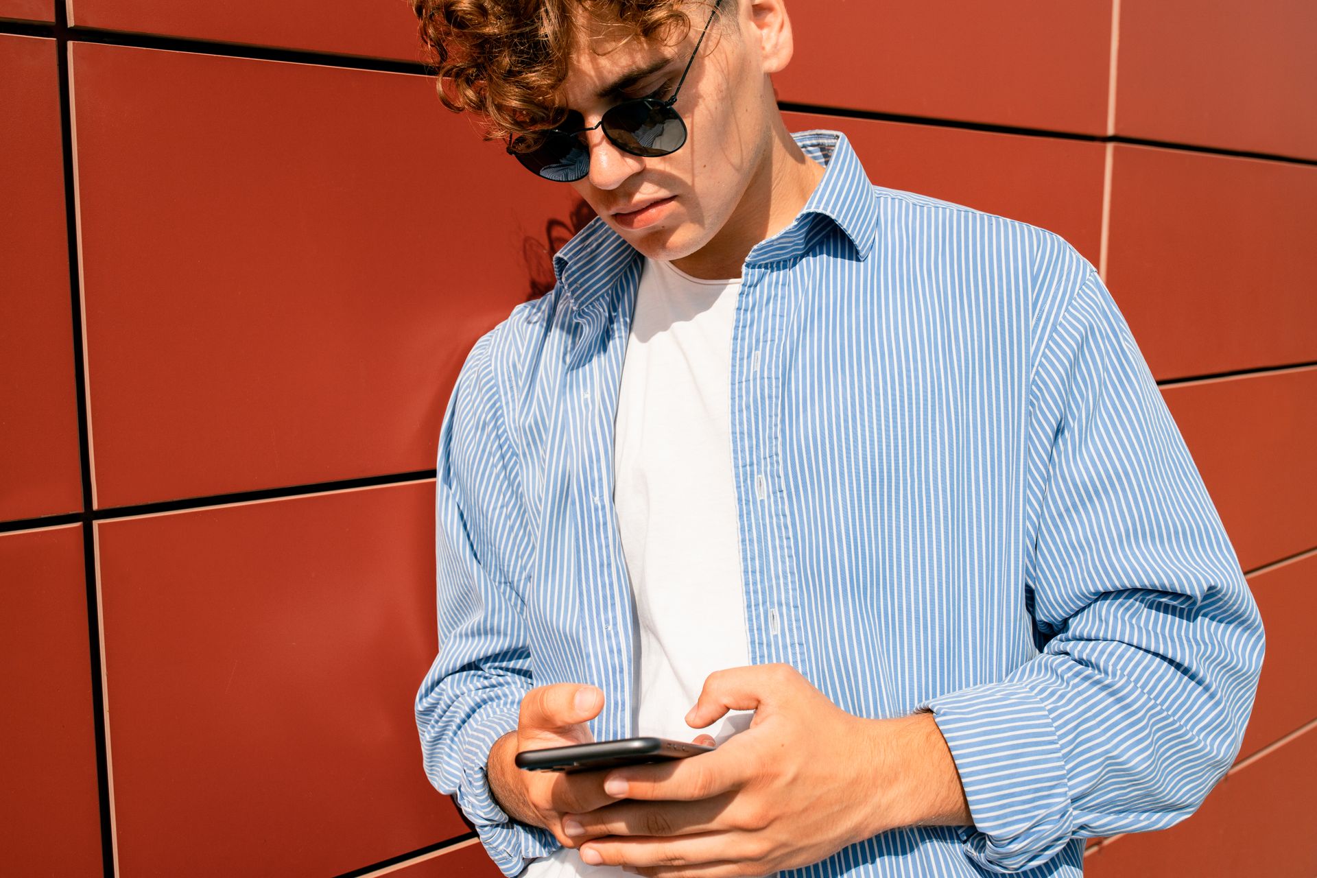 Confident young man wearing sunglasses and striped shirt messaging on his smartphone outdoors.