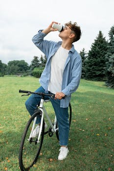Young man in casual attire hydrates while seated on bicycle in a park.