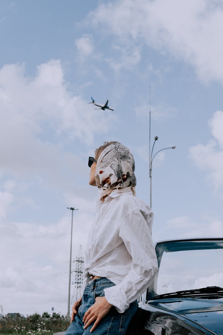 A Stylish Woman Leaning On A Car Looking At A Plane In The Sky