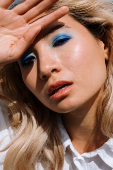 Close-up portrait of a woman with blonde hair and vibrant blue eyeshadow, hand shielding face.
