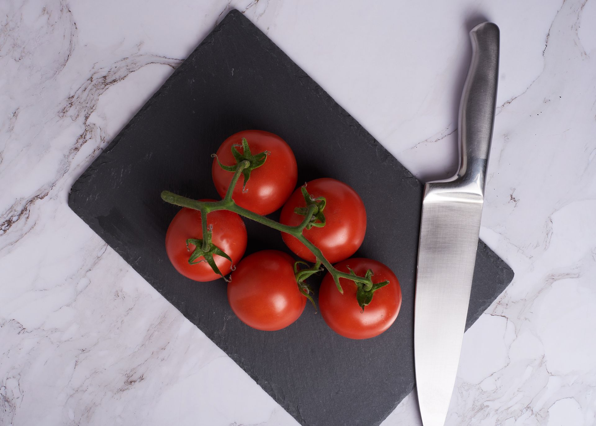 Top view of vine tomatoes on a slate cutting board with a stainless steel knife on a marble counter.