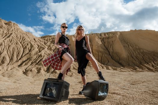 Two women in fashionable attire stand in a desert landscape with vintage televisions, striking a bold pose.