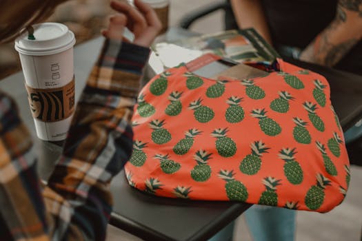 Colorful pineapple-patterned bag on a table with coffee cup, books, and people conversing.